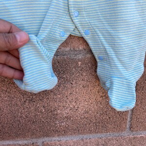 Puede incluir: Mono de beb&eacute; a rayas azules y amarillas con una aplicaci&oacute;n de elefante azul y naranja y el texto "John Lennon" bordado en la parte delantera. El mono tiene broches en la entrepierna y los hombros.