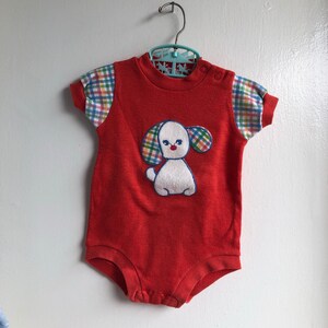 Puede incluir: Unisie rojo para beb&eacute; con una aplicaci&oacute;n de cachorro blanco. El cachorro tiene orejas de cuadros azules, verdes y amarillos y una nariz roja. El unisie tiene mangas cortas con tela de cuadros en las mangas.