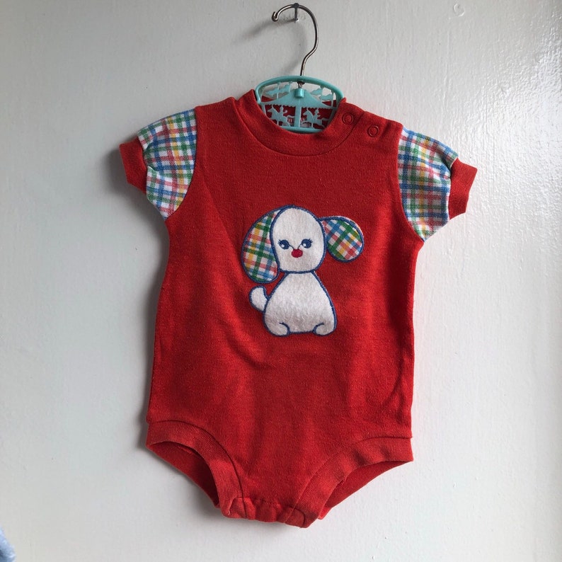 Puede incluir: Unisie rojo para beb&eacute; con una aplicaci&oacute;n de cachorro blanco. El cachorro tiene orejas de cuadros azules, verdes y amarillos y una nariz roja. El unisie tiene mangas cortas con tela de cuadros en las mangas.