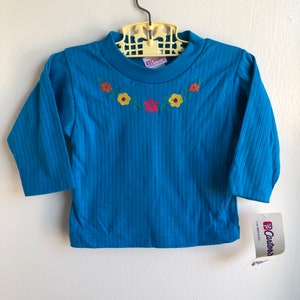 May include: A blue long-sleeved shirt with a ribbed texture and embroidered flowers in orange, yellow, red, and green. The shirt has a crew neck and is labelled "Carter's".