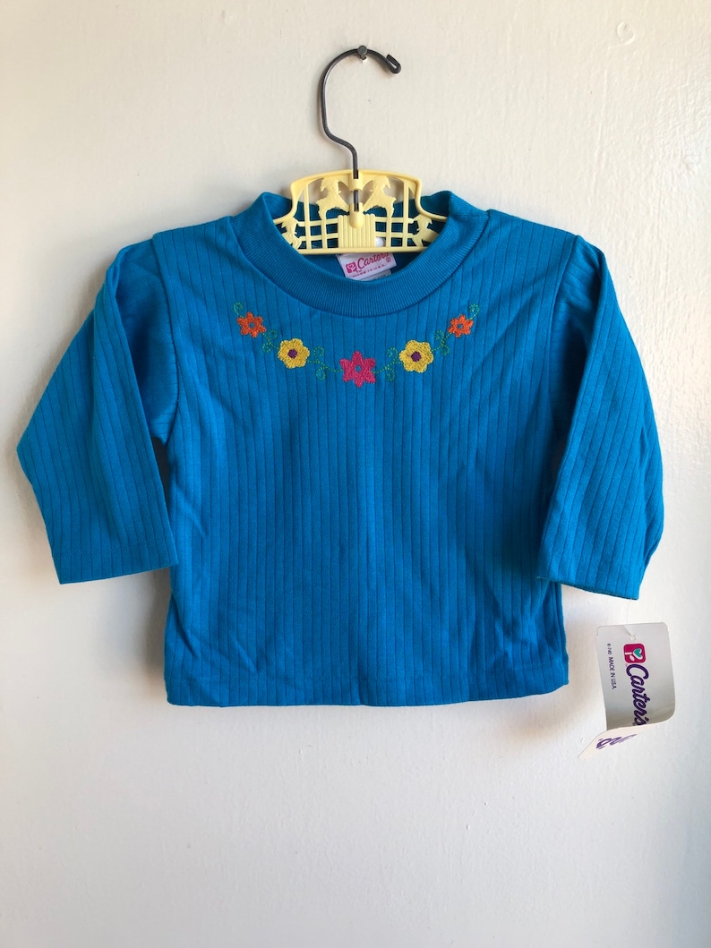 May include: A blue long-sleeved shirt with a ribbed texture and embroidered flowers in orange, yellow, red, and green. The shirt has a crew neck and is labelled "Carter's".