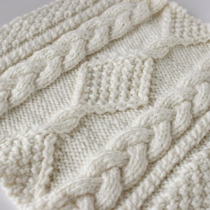 May include: A white knitted fabric with a cable knit pattern. The fabric has a textured surface and a subtle sheen.