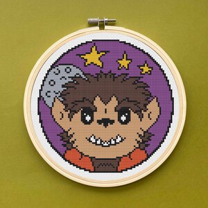 May include: Cross-stitch embroidery of a cartoon werewolf in a wooden hoop. The werewolf has brown fur, a tan face, and orange clothing. The background is purple with yellow stars and a gray moon.