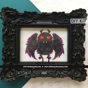 Könnte beinhalten: Ein schwarz gerahmtes Kreuzstich-DIY-Kit mit einem pixeligen, mottenartigen Wesen mit roten Augen und lila Flügeln. Der Rahmen hat aufwendige Details. Der Text "DIY KIT" ist sichtbar. Die Website spotyourcolors.com ist ebenfalls sichtbar.