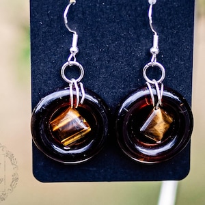 May include: A pair of brown glass earrings with a gold-toned square stone in the center. The earrings are hanging from silver hooks and are labeled "Handmade with love".