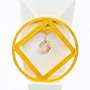 May include: A gold-colored pendant featuring a circle and a square design, with a smaller square pendant hanging from it. The pendant is set against a white background. The design is geometric and modern.