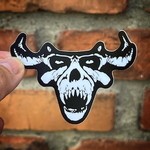 May include: Black and white sticker of a skull with horns and a large, open mouth with sharp teeth. The skull is in a menacing expression.