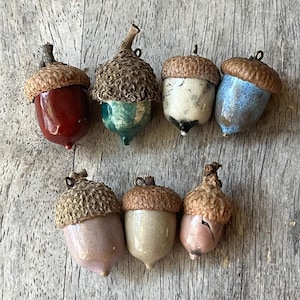 May include: A collection of seven ceramic acorn ornaments. The acorns feature a variety of colors, including red, green, white, blue, pink, and beige. Each acorn has a textured cap and a small loop for hanging.