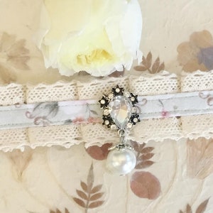 May include: A delicate white lace choker with a light blue ribbon and floral print. The ribbon features a silver charm with a clear rhinestone and a pearl drop.