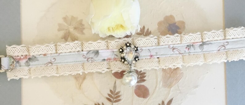 May include: A delicate white lace choker with a light blue ribbon and floral print. The ribbon features a silver charm with a clear rhinestone and a pearl drop.