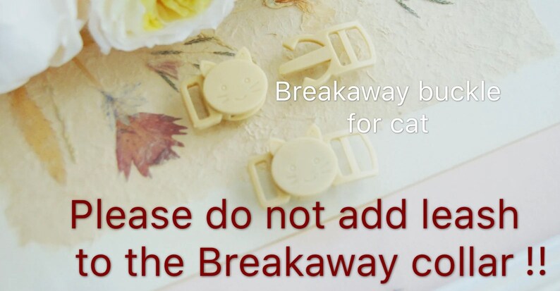 May include: Two beige plastic breakaway buckles for cat collars. The buckles have a cat face design and the text "Breakaway buckle for cat" is written on the image. The text "Please do not add leash to the Breakaway collar!" is written in red.