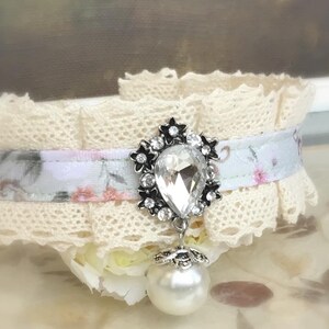May include: A delicate white lace choker with a floral ribbon and a crystal pendant. The pendant is a teardrop shape with a silver setting and a pearl drop.