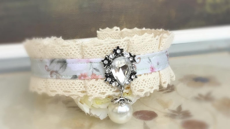 May include: A delicate white lace choker with a floral ribbon and a crystal pendant. The pendant is a teardrop shape with a silver setting and a pearl drop.