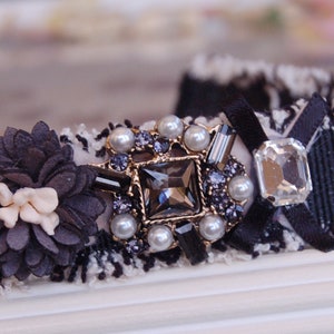 Può includere: Una fascia per capelli in tweed bianco e nero con un accento floreale decorativo e un abbellimento centrale gioiello. Il fulcro presenta un grande cristallo quadrato circondato da perle e cristalli più piccoli, con un fiocco di nastro nero.
