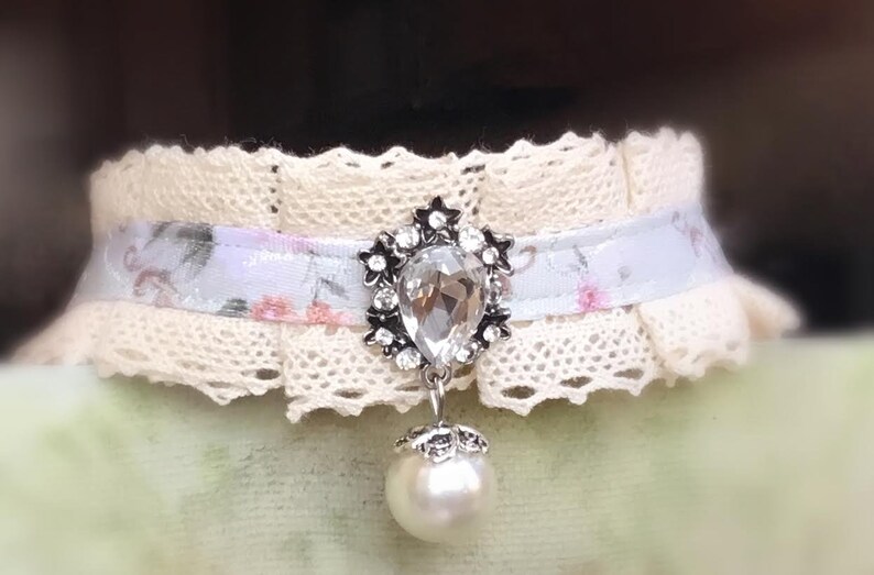 May include: A delicate white lace choker with a floral ribbon and a dangling pearl and crystal pendant.