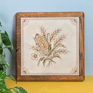 May include: Square wooden trivet with a ceramic tile insert. The tile features a detailed illustration of corn, wheat, and flowers in shades of tan, green, and brown. The trivet has a light brown wooden frame.