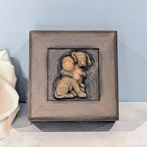 May include: A square, dark gray wooden frame containing a relief sculpture of a person embracing a dog. The sculpture is rendered in muted tan and brown tones, set against a dark, textured background within the center of the frame.