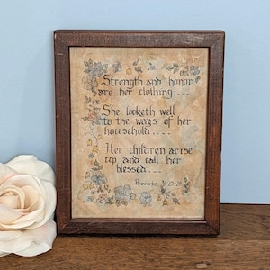 May include: A framed print with a dark brown wooden frame. The print features calligraphy with the text "Strength and honor are her clothing..." and floral illustrations in shades of blue and beige. The text also includes "Proverbs 31:25-28".