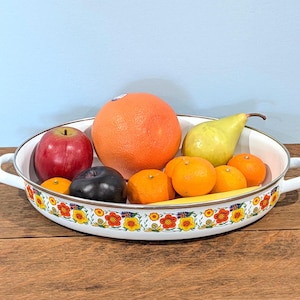 May include: An oval white enamel serving dish with two side handles, decorated with a colorful floral pattern of orange and yellow flowers. The dish is filled with a variety of fresh fruit, including a large grapefruit, a red apple, a green pear, and several small oranges.