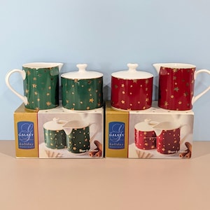 May include: Two sets of Galaxy Holiday Dinnerware sugar and creamer containers. One set is dark green and the other is red, both featuring gold star patterns. Each set includes a lidded sugar bowl and a creamer pitcher, displayed on their boxes.