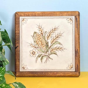 May include: A square ceramic tile framed in dark wood featuring a central illustration of a corn cob surrounded by wheat stalks and small flowers. The tile has a cream-colored background with a decorative floral motif in each of the four corners.