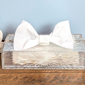May include: A rectangular clear glass tissue box cover featuring a sunburst pattern of etched lines radiating from the center. A single white paper tissue is pulled through the top opening, folded into a bow shape that sits atop the glass surface.