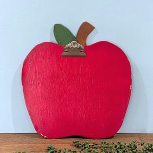 May include: A red apple-shaped wooden clipboard with a green leaf and brown stem. The clipboard has a silver metal clip at the top. The apple is painted red with visible wood grain and white distressed edges.