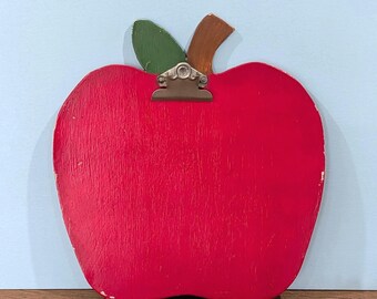 May include: A red apple-shaped wooden clipboard featuring a green leaf and a brown stem at the top. A silver metal clip is attached to the center of the apple, designed to hold papers securely. The surface of the wood shows a visible grain texture.