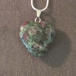 May include: A heart-shaped pendant with a silver chain. The pendant is made of a green stone with red and black inclusions. The pendant is attached to a silver bail.