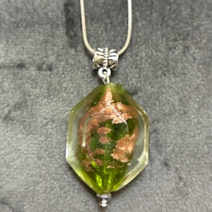 May include: A silver chain necklace with a pendant. The pendant is a green and copper-colored glass gem with a geometric shape. The gem is suspended from a silver bail and chain. The background is a gray fabric.
