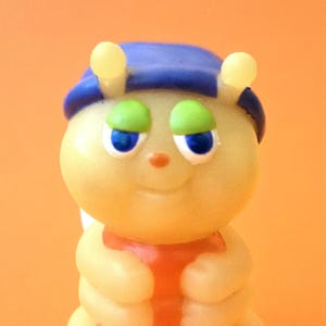 May include: A vintage, smiling caterpillar figurine. The toy is yellow with a blue hat, green eyelids, and an orange belly. The caterpillar's arms are crossed, and it has two yellow antennae.