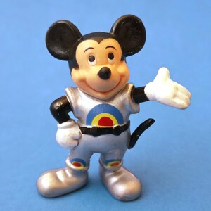 Peut inclure: Figurine vintage de Mickey Mouse dans une combinaison spatiale argent&eacute;e avec un motif de cible sur la poitrine. La figurine a des oreilles noires, un visage souriant et des gants blancs. Le fond est bleu uni.