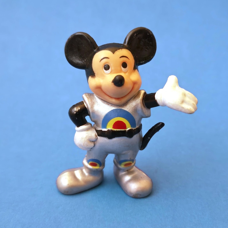 Peut inclure: Figurine vintage de Mickey Mouse dans une combinaison spatiale argent&eacute;e avec un motif de cible sur la poitrine. La figurine a des oreilles noires, un visage souriant et des gants blancs. Le fond est bleu uni.