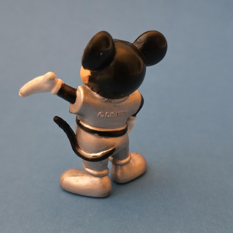 Peut inclure: Petite figurine de Mickey Mouse argent&eacute; avec des oreilles noires et une queue noire. La figurine porte une combinaison spatiale argent&eacute;e avec des d&eacute;tails noirs et des gants blancs. Le mot "&copy; DISNEY" est imprim&eacute; au dos.