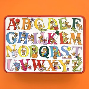 May include: A rectangular, vintage alphabet tin with a red border. Each letter of the alphabet is displayed in a different color, with a teddy bear illustration. The background is white, and the tin is set against an orange background.