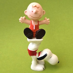 May include: A figurine of Charlie Brown sitting atop Snoopy. Charlie Brown is wearing an orange outfit with the number 1. Snoopy is white with black ears and a red collar. The background is a solid green.