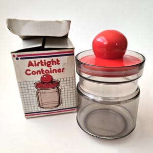 May include: A clear glass airtight container with a red lid and knob, next to its original box. The box has the words "Airtight Container" and a drawing of the jar. The jar is empty.