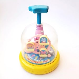 May include: A yellow and pink toy merry-go-round with a clear dome. The toy has a blue star on top and a push button on the bottom that says "Push 'n Merry-Go-Round" and "Tomy".