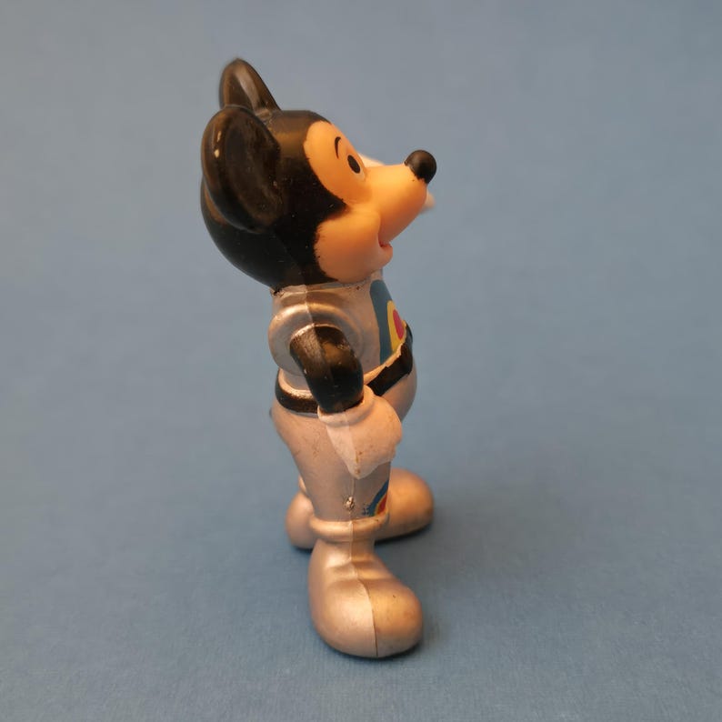 Peut inclure: Petite figurine vintage de Mickey Mouse en combinaison spatiale. La figurine est principalement argent&eacute;e avec des oreilles noires et un nez noir. La combinaison spatiale pr&eacute;sente un motif circulaire bleu et rouge sur la poitrine. La figurine est pos&eacute;e sur une surface bleue.