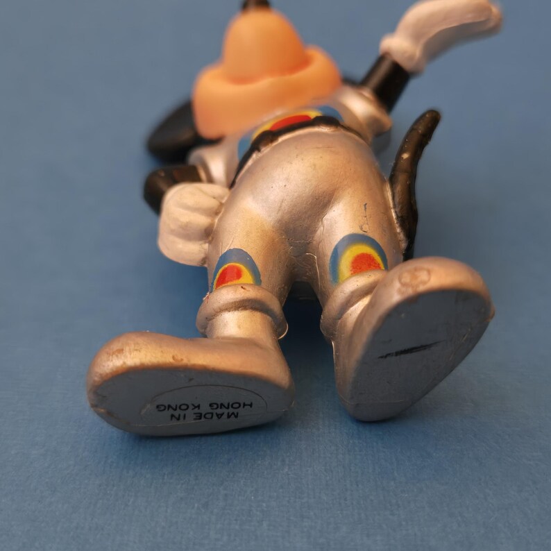 Peut inclure: Figurine de Dingo argent&eacute; avec un chapeau et un nez beiges, des oreilles noires et des gants blancs. La figurine pr&eacute;sente des motifs circulaires color&eacute;s sur les jambes et porte la mention "MADE IN HONG KONG" sur le pied. Sur fond bleu.