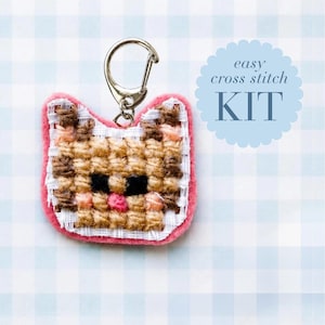 May include: A cat-shaped cross stitch keychain featuring tan, brown, and black thread on white fabric with a pink felt border and a silver metal lobster clasp. The text "easy cross stitch KIT" is printed in blue on a scalloped circular background.
