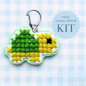 May include: A turtle-shaped cross stitch project featuring a green shell and yellow body with a black eye, attached to a silver metal lobster clasp keychain. The text "easy cross stitch KIT" is printed in a blue scalloped circular graphic.