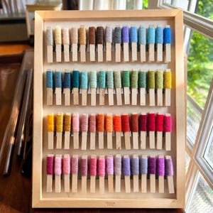 May include: A wooden display frame filled with a variety of yarn samples. Each sample is wound around a small wooden stick. The colours include a spectrum of shades, from soft pastels to bold hues. The frame is made of light-coloured wood, and the yarn is arranged in rows.