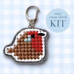 May include: A cross stitch keychain featuring a bird with a red chest and brown wings, attached to a silver metal lobster clasp. The text "easy cross stitch KIT" is printed in a blue scalloped circle next to the bird-shaped embroidered piece.