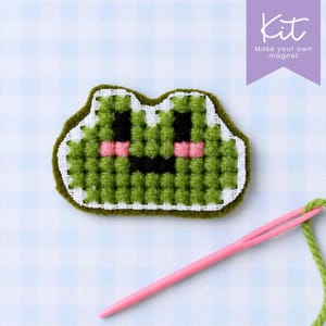 Pode incluir: Um kit de íman de rã verde pixelizada com agulha rosa e fio verde. A rã tem olhos e boca pretos e bochechas rosa. O kit inclui o texto "Kit" e "Make your own magnet" numa faixa roxa.