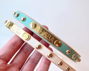 May include: Two leather collars featuring gold-tone metal studs and rectangular nameplates. One collar is teal with the name SHEBA engraved, and the other is light beige with the name SHEBA and the phone number 123-456-7890 engraved on the plate.