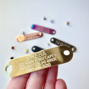 May include: A gold-coloured metal pet tag with the name "SHEBA" and address details engraved in white. Other colourful tags are scattered in the background. The tag has two holes for attachment.