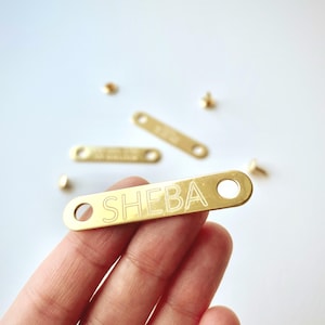 May include: A gold-colored metal tag with the name "SHEBA" engraved on it. The tag has two holes and is held by a hand. Several other similar tags and small gold rivets are scattered on a white surface.