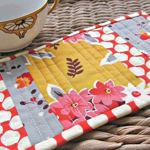 May include: A rectangular quilted fabric coaster featuring a patchwork design of mustard yellow, grey, and red floral patterns. The piece is constructed with straight-line stitching and finished with a red and white patterned fabric border.