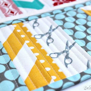 May include: A quilted fabric organizer featuring a patchwork design with light blue and gray polka dots, mustard yellow diagonal stripes, and a white panel with gray scissor icons. The piece is stitched with vertical lines and includes the text Saltwater Quilts.